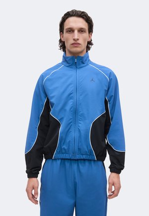 Man with medium-length dark hair wearing a blue and black zip-up sports jacket and matching blue pants standing against a plain background.