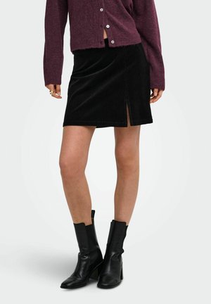 Femme portant une mini-jupe noire en velours côtelé avec une fente devant, un pull violet boutonné et des bottines noires à talons.