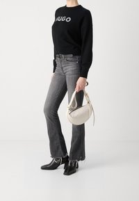 Schwarzer Pullover mit "HUGO" Schriftzug, graue Schlagjeans mit fransengesäumtem Saum, schwarze spitz zulaufende Ankle Boots und eine cremefarbene, geschwungene Handtasche.
