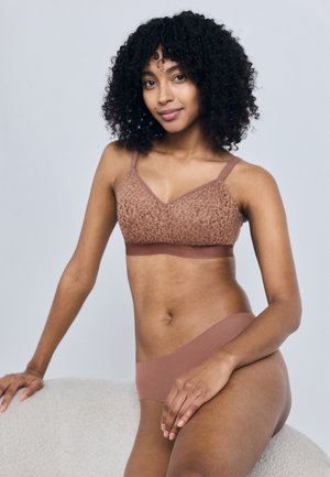 Vrouw met krullend haar, gekleed in een bijpassende bruine kanten beha en naadloos ondergoed, leunend op een witte textuurige ondergrond tegen een lichtgrijze achtergrond.