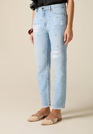 Lyseblå slidte jeans med perlestuds, båret med beige spidse flade sko med ormemønstrede stropper og en sort top, delvist synlig.
