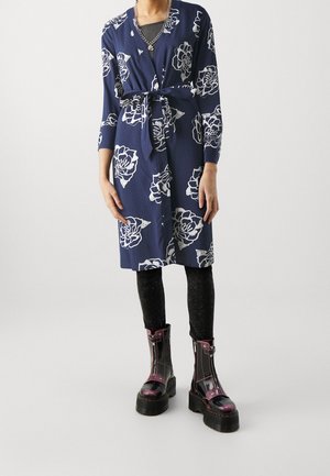 Personne portant une robe bleu marine longueur genou avec des motifs floraux blancs, des leggings noirs, des bottes épaisses à plateforme et un collier chaîne argentée.