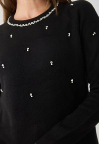 Fekete kötött pulóver kerek nyakkal, az elején szétszórt műgyöngyökkel és strasszokkal díszítve, texturált megjelenést kölcsönözve.