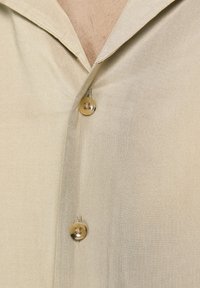 Camisa de un tono beige claro con una textura sedosa, que presenta un cuello con muescas y botones dorados, destacando un diseño suave y aerodinámico.