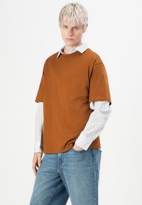 Brun T-shirt med korte ærmer lagd over en hvid stribet langærmet skjorte. Kombineret med lyseblå jeans. Simpelt design og pasform.