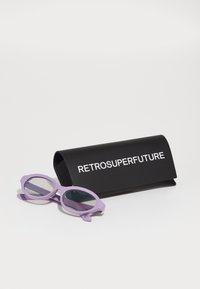 Gafas de sol moradas con un diseño angular único y lentes transparentes, junto a un estuche negro con "RETROSUPERFUTURE" en texto blanco.