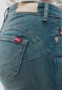 Mėlynos džinsinės kelnės su tekstūruotu paviršiumi. Ypatybės apima prekės ženklo etiketę ant juosmens ir mažą raudoną etiketę ant priekinio kišenės.