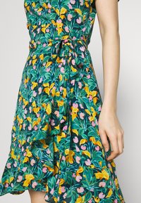 Robe portefeuille fleurie avec des motifs verts, jaunes et roses. Comprend des manches courtes, un ourlet volanté et une ceinture en tissu à la taille.