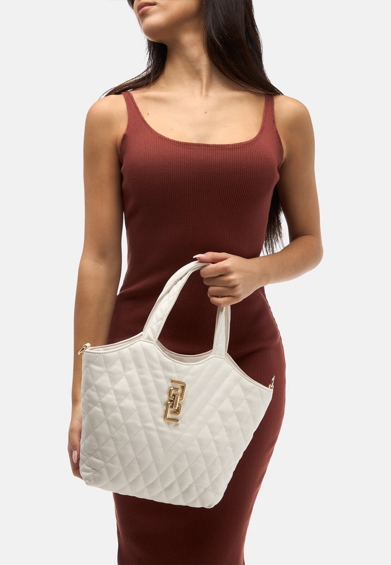 Femme en robe marron sans manches tenant un sac à main blanc matelassé avec des détails dorés sur un fond uni.