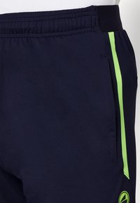 Shorts de sport bleu marine avec une taille élastique, des poches latérales et des rayures néon vertes contrastantes sur les côtés. Texture du tissu lisse.