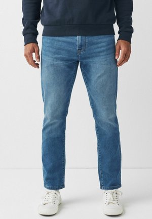 Mann trägt mittelblaue Jeans mit geradem Schnitt, weiße Sneaker und ein dunkles Langarmshirt, steht mit dem Gesicht nach vorne vor einem einfarbigen Hintergrund.