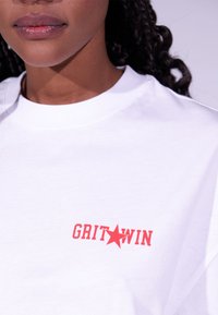 Bílý bavlněný tričko s červeným textem "GRIT WIN" a hvězdnou grafikou, standardní design s kulatým výstřihem, hladká textura.