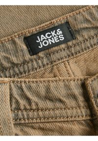 Nærbillede af brun denimstof med detaljeret syning og en sort mærkat med teksten "JACK & JONES" syet på materialet.
