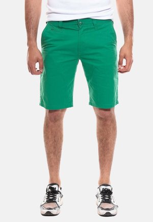 Shorts en coton verts avec une fermeture éclair et un bouton, dotés de poches et d'une coupe droite. Portés avec des baskets noir et blanc.