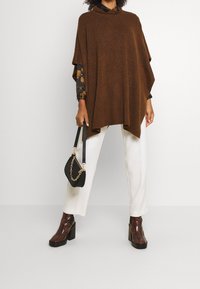Poncho marrone lavorato a maglia con collo alto, abbinato a una blusa a maniche lunghe a fiori scuri, pantaloni bianchi e stivali a piattaforma lucidi marroni.