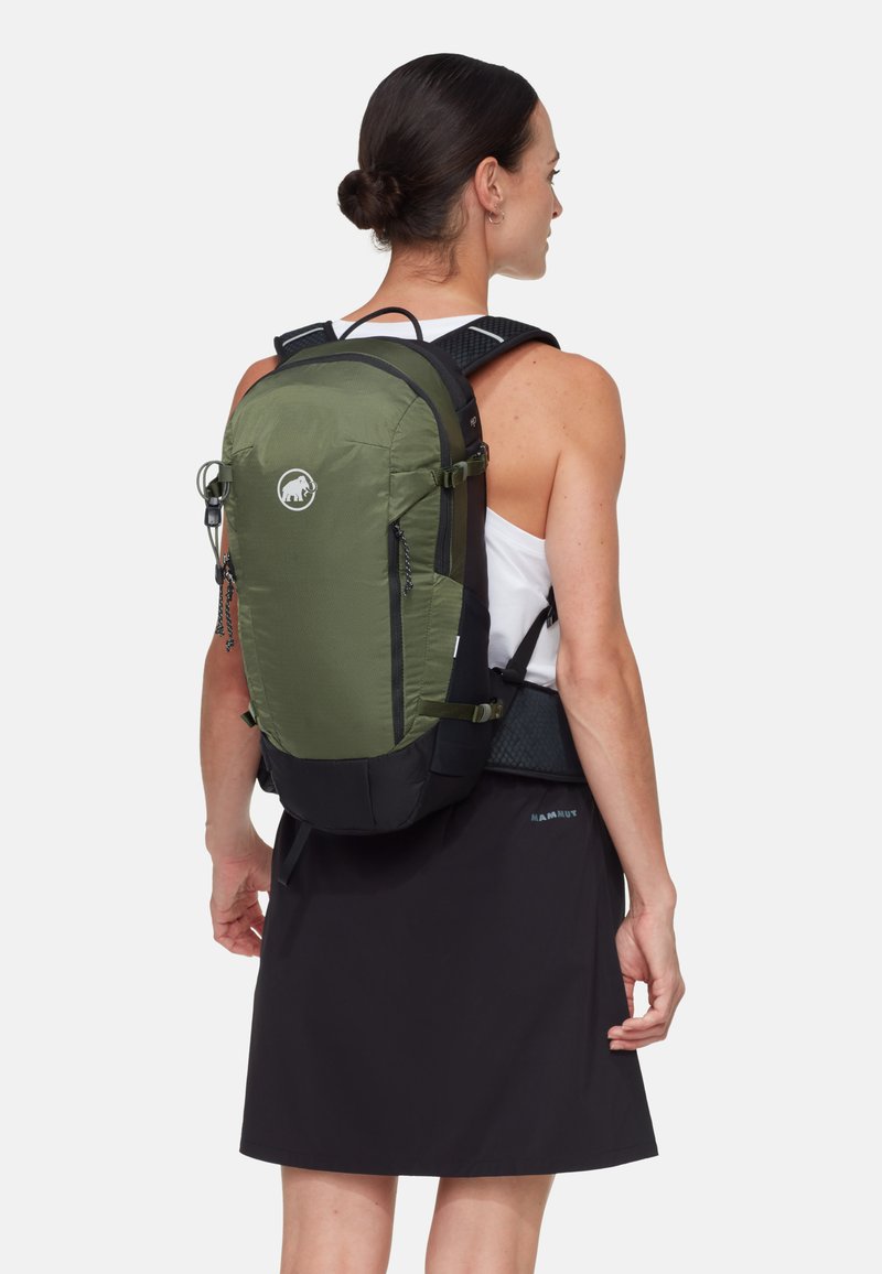 Frau mit dunklem Haar im Dutt, trägt ein weißes Tanktop und einen schwarzen Rock, trägt einen grünen und schwarzen Rucksack mit einem Logo vor einfarbigem Hintergrund.