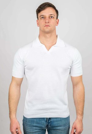 Uomo in piedi di fronte, che indossa una polo bianca semplice a maniche corte e jeans blu su uno sfondo grigio chiaro.