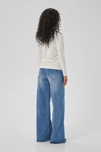 Woman with curly hair wearing a white long-sleeve top and wide-leg blue jeans, standing facing away on a plain gray background.