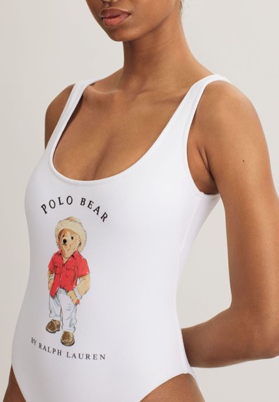 Vit baddräkt med motiv av en björn i röd skjorta, med texten "POLO BEAR BY RALPH LAUREN" nedanför. Mjuk textur.