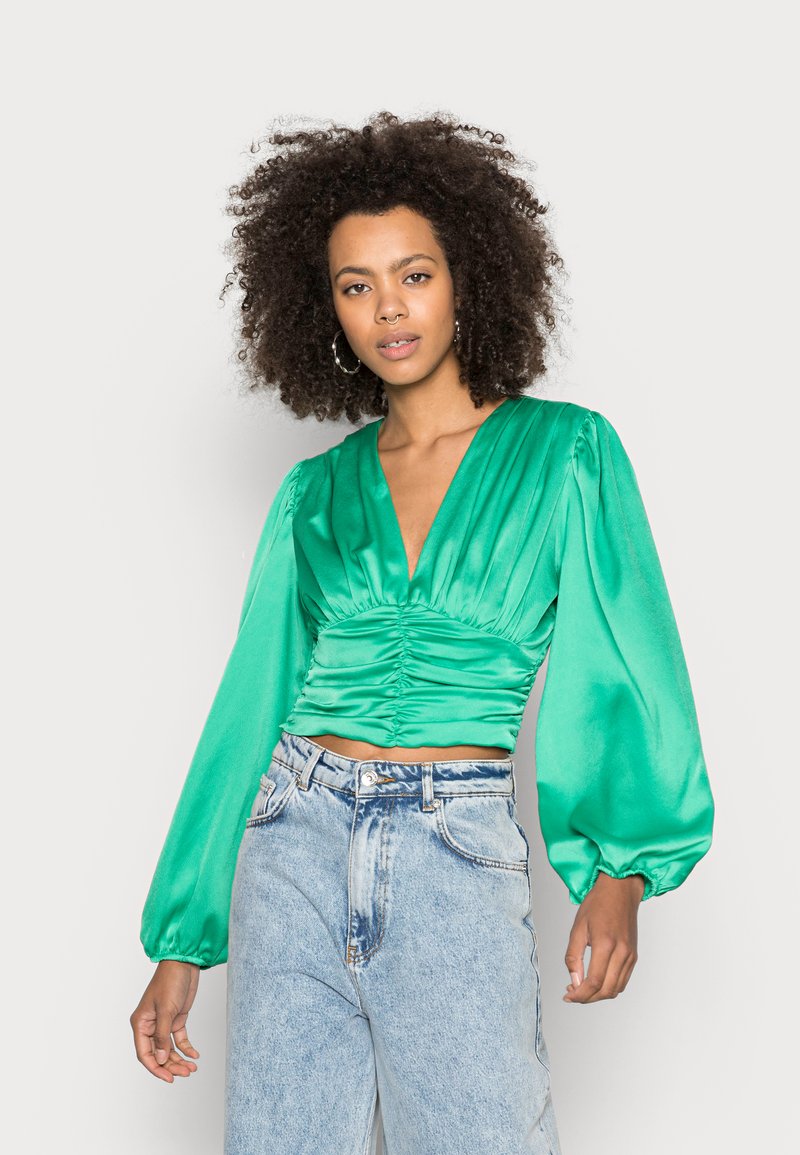 Blusa de satin verde com mangas longas bufantes, decote em V profundo e detalhes franzidos na cintura. Combinada com jeans de cintura alta em denim claro.