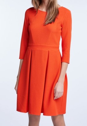 Femme portant une robe texturée orange vif avec des manches trois quarts et une taille ajustée, debout devant un fond clair uni.