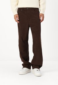 Pantalones de pana marrón con un corte relajado, que presentan un cierre de botón y cremallera, bolsillos frontales y piernas anchas, combinados con zapatillas blancas.
