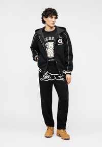 Μαύρο μπουφάν με φερμουάρ και λευκές λεπτομέρειες, πάνω από μαύρο γραφικό μπλουζάκι και μαύρη φόρμα με λευκό σχέδιο γάτας. Καφέ παπούτσια.