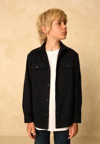Jachetă neagră cu două buzunare pe piept, închidere cu nasturi și mâneci lungi. Purtați deasupra unui tricou alb și asortată cu blugi închisi.