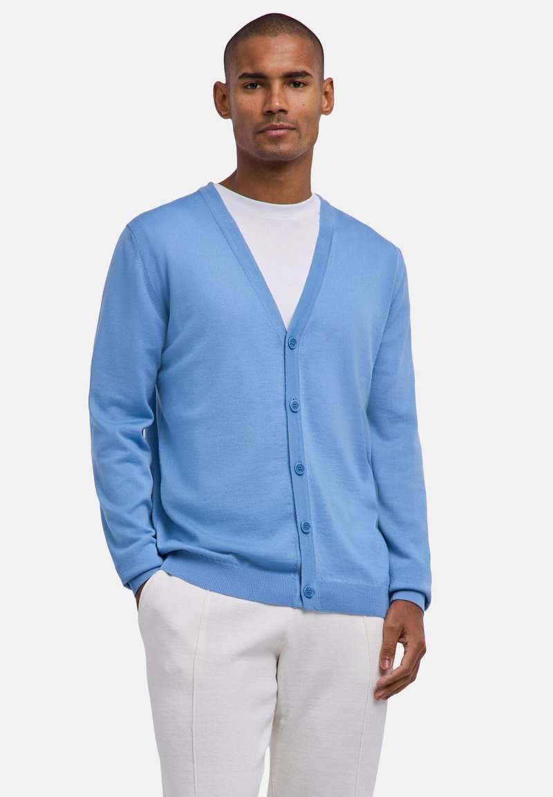 Homme portant un cardigan bleu clair boutonné sur une chemise blanche et un pantalon clair, debout avec une main dans la poche.