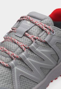 Šedá atletická topánka Columbia s červenými akcentmi na šnúrkach, vrchnou časťou z sieťoviny, prešitými prekryvmi a detailom červenej polstrované golierovej časti.