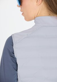 Ljusgrå vadderad väst med vertikal quiltning och hög krage, lager på lager över en blå långärmad tröja. Slät textur och figursydd design.