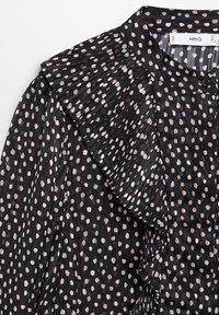 Blouse noire avec des pois irréguliers rose clair, dotée d'un détail volanté plissé le long de l'épaule et à l'avant.