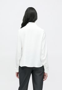 Balta krekla ar garām piedurknēm, rotāta ar ruļļu apkakli un pogātām aprocēm, izgatavota no teksturēta materiāla. Valdāts kopā ar tumšām biksēm.