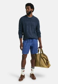 Maglione a maglia navy con motivo testurizzato, pantaloni corti blu, calzature marroni e borsa da viaggio beige, con calzini a contrasto con scritte rosse.