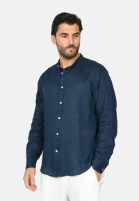 Camicia in lino blu navy con colletto alla mandarino, chiusura frontale con bottoni e maniche lunghe. Tessuto morbido, vestibilità casual, indossata da un modello.