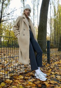 Abrigo teddy beige con diseño de doble botonadura, combinado con pantalones oscuros y zapatillas New Balance plateadas, en un entorno otoñal.