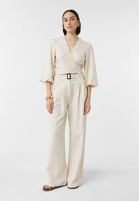Helles, wickelartiges Blusenshirt mit Puffärmeln, kombiniert mit hochTaillierten, weit geschnittenen Hosen. Enthält einen Gürtel und flache Sandalen.