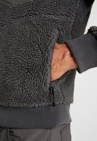 Szary polarowy sweter z teksturowanym, puszystym materiałem. Posiada kieszeń na zamek i ściągacze na mankietach. Krój jest luźny i ciepły.