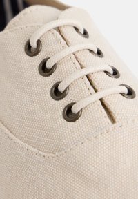 Primer plano de una zapatilla de lona beige que presenta ojales de metal redondos y cordones de color claro sobre una superficie texturizada.