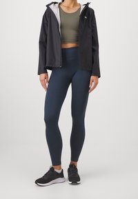 Chaqueta ligera negra, top corto verde oliva y leggings azul marino de cintura alta combinados con zapatos deportivos negros. Textura suave, diseño ajustado.