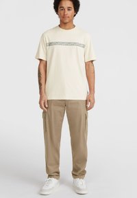 Beige katoenen t-shirt met een horizontale patroonaccent in het midden, gecombineerd met kaki cargo shorts en witte sneakers.