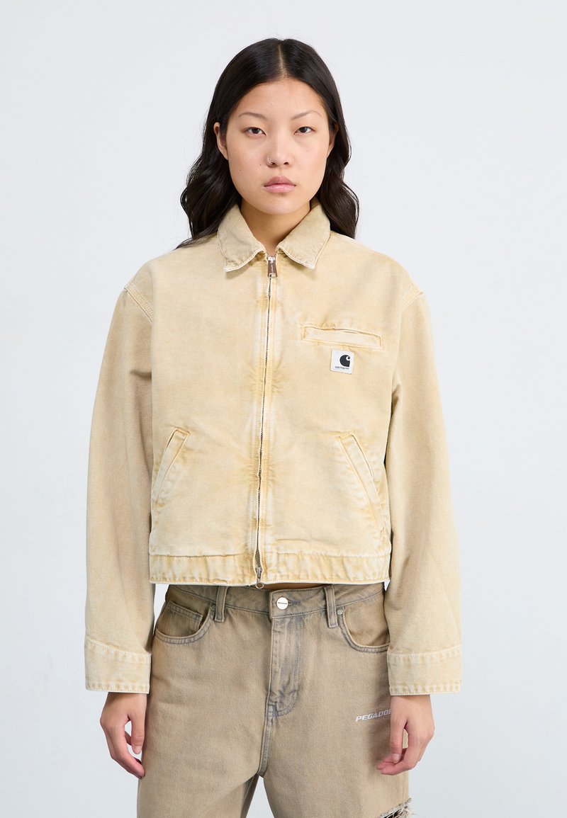 Veste en denim beige clair avec une fermeture éclair à l'avant, un col pointu et deux grandes poches avant. Présente un petit patch logo sur la poitrine.