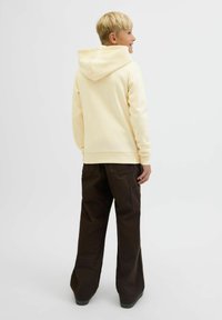 Svetlolasi otrok se smeji, stoji in se obrne bočno, oblečen v kremni kapuco in ohlapne temno rjave cargo hlače pred enostavno ozadje.