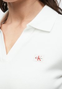 Biely žebrovaný polokošeľa s golierovým výstrihom. Na ľavej strane je červeno-biely nášivka s logom Calvin Klein.