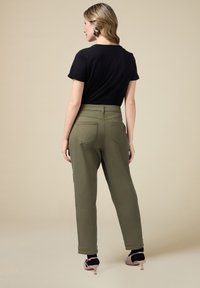 Schwarzes Baumwoll-T-Shirt kombiniert mit olivgrünen, schmal geschnittenen Hosen. Die Hosen verfügen über Gesäßtaschen und Bündchen an den Knöcheln. Neutrale Hintergrund.