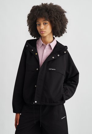 Giacca antivento nera con cappuccio, bottoni a pressione, tasche frontali e logo brandizzato. Indossata sopra una camicia rosa chiaro lavorata a trama.