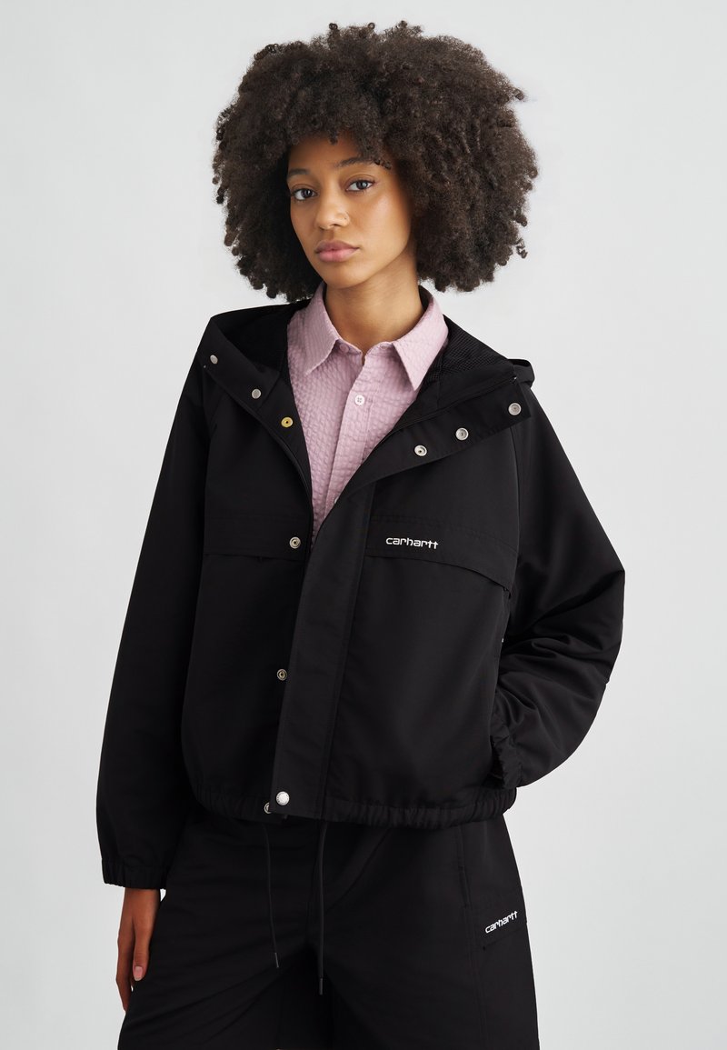 Giacca antivento nera con cappuccio, bottoni a pressione, tasche frontali e logo brandizzato. Indossata sopra una camicia rosa chiaro lavorata a trama.