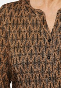 Braun gemustertes Hemd mit einem geometrischen schwarzen Design und Baummotiven; hat einen runden Ausschnitt und einen Knopfverschluss.