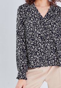 Blouse noire à manches longues avec un motif floral blanc, dotée d'un col en V et de poignets à volants texturés. Confectionnée en tissu léger.