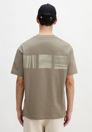 Mann trägt taupefarbenes T-Shirt mit hellen grafischen Quadraten und Text "Marc O’Polo" am oberen Rücken sowie schwarze Baseballkappe, Blick nach hinten gewandt.
